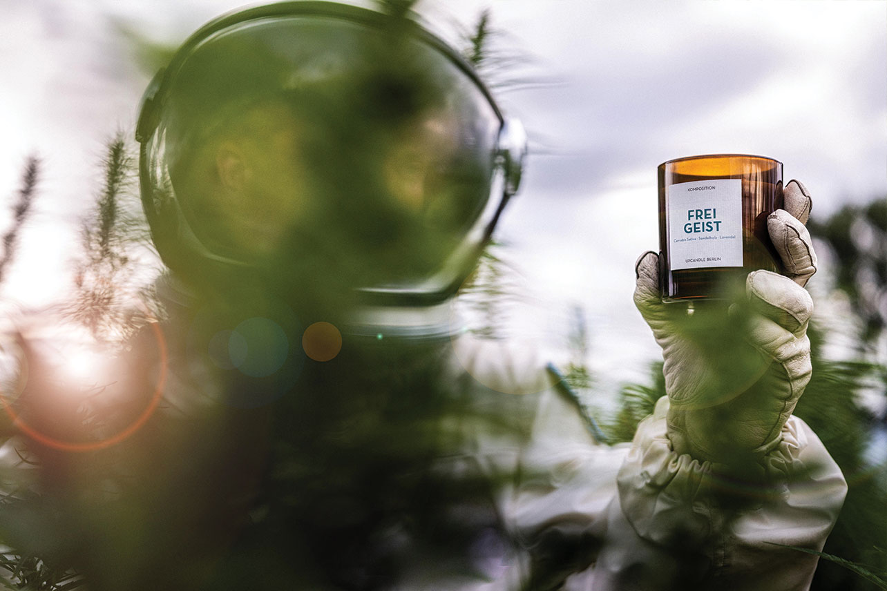 Hand des Astronauten zeigt UpCandle® Duftkerze im grünen Hanffeld bei Produktshooting
