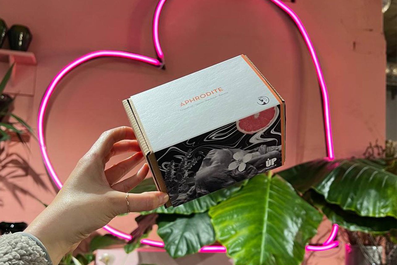 Hand hält APHRODITE Kerzen-Packaging mit Neon-Herz und Blumen – UpCandles neue sinnliche HAPPINESS-Duftkerze