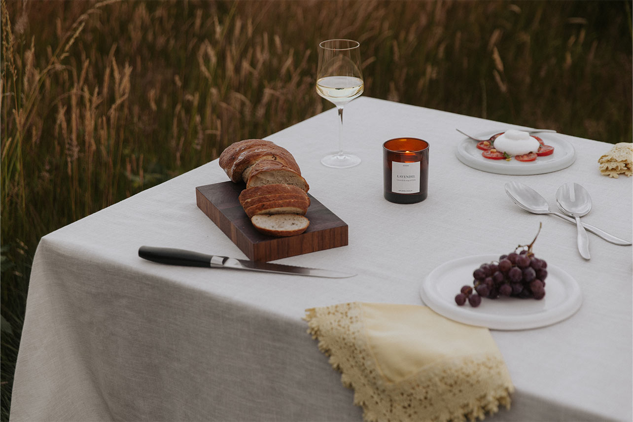 Gedeckter Tisch im Spätsommer mit Brot, Wein und der brennenden UpCandle Aromakerze Lavendel mit Holzdocht vor einem reifen Kornfeld.
