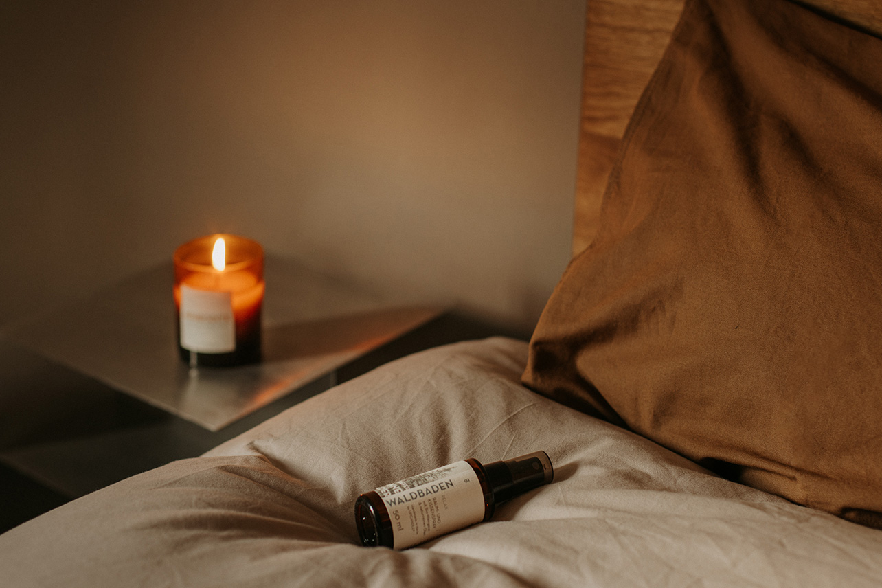 Das UpCandle Raumspray Waldbaden liegt auf einer gemütlichen Bettdecke, im Hintergrund ein Nachttisch mit einer brennenden Aromakerze in erdigen, braunen Farbtönen.